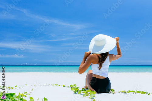 A view from the back of a girl in a white straw hat sitting on the shore of a tropical beach with white sand and bruise color of the sea