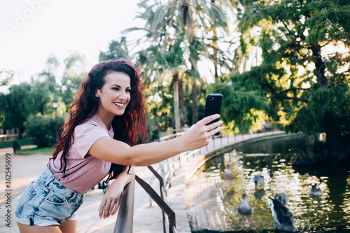 Beautiful brunette curly female influencer posing for photo on mobile phone resting in city park with lake on weekends,charming female 20s using smartphone for making selfie on free time vacations #362545699