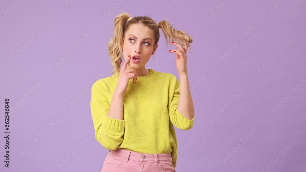 An unrivaled blonde girl thoughtfully puts hair on her finger, shows the index finger of her hand near her cheek in an isolated studio on a purple background An unrivaled blonde girl thoughtfully puts hair on her finger, shows the index finger of her hand near her cheek in an isolated studio on a purple background