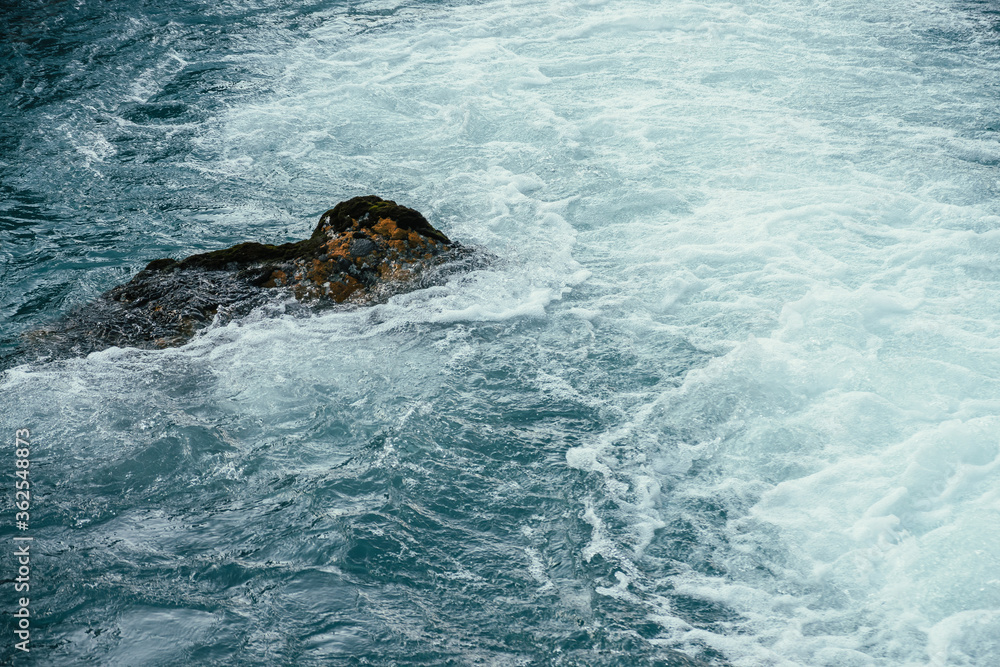 Fototapeta premium Mossy stone in azure water of mountain river close-up. Blue nature background with boulder with moss in turquoise water of mountain creek. Full frame of sea surf. Stone in ocean surf. Backdrop of tide