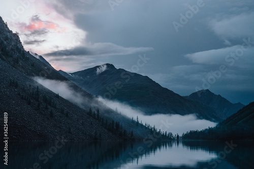 Low cloud above highland lake. Silhouettes of trees on hillside along mountain lake in dense fog. Reflex of pines to calm water. Alpine tranquil landscape at early morning. Ghostly atmospheric scenery