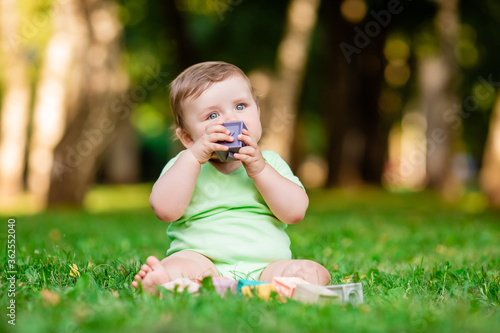 Cute kid in a green bodysuit sitting on the grass in summer plays with developing toys. The baby gnaws silicone cubes. Child development up to one year