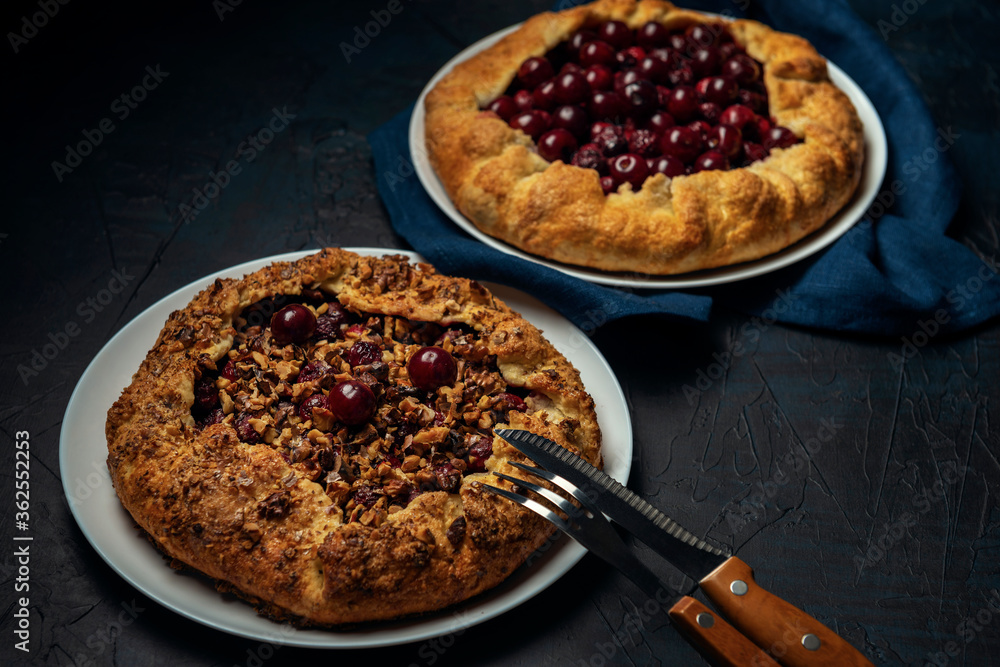 Two galettes with ripe red cherry and walnut filling on dark blue background. Homemade sweet open pie with napkin and cutlery. Two galettes with ripe red cherry and walnut filling on dark blue background. Homemade sweet open pie with napkin and cutlery.