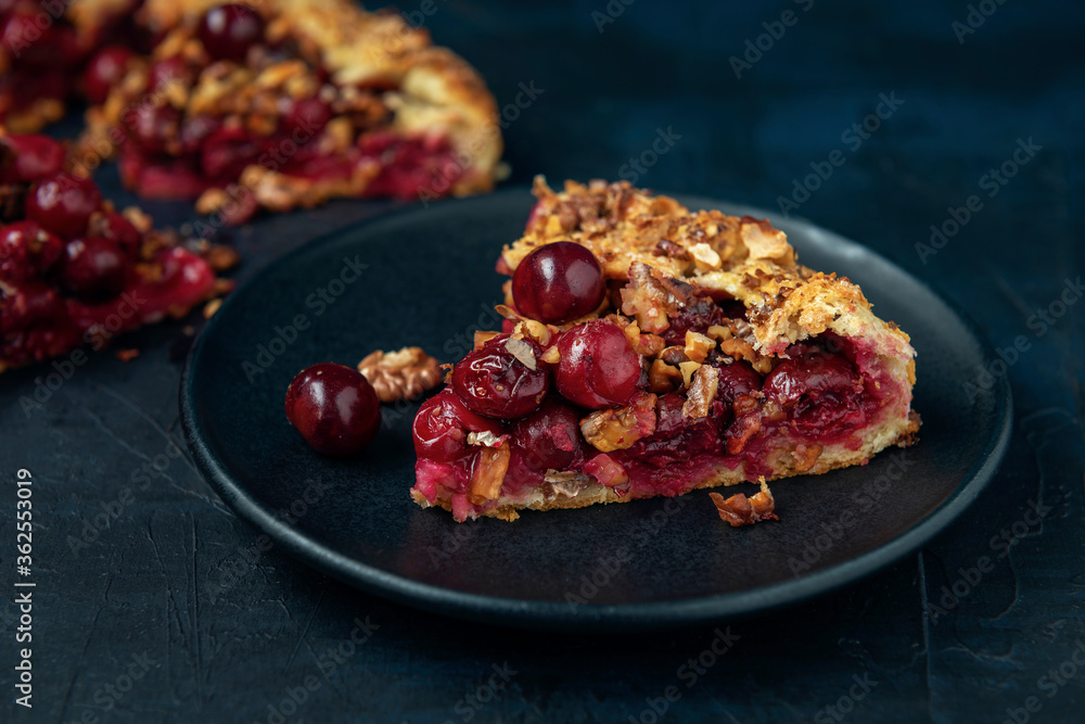 A piece of galette with ripe red cherry filling on black plate. Homemade sweet open pie on dark textured backdrop. A piece of galette with ripe red cherry filling on black plate. Homemade sweet open pie on dark textured backdrop.