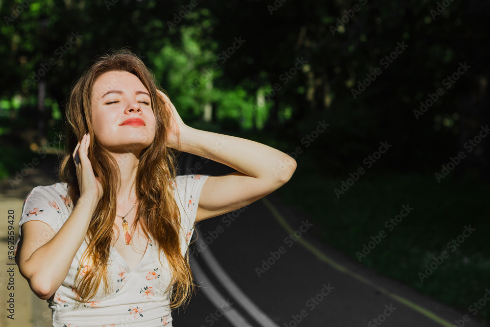 Naklejka premium Portrait young woman in white top touching her hair and enjoying sun light on the drive way in the park . Solo outdoor activity. End quarantine. Alone teenage girl in the nature. Close up