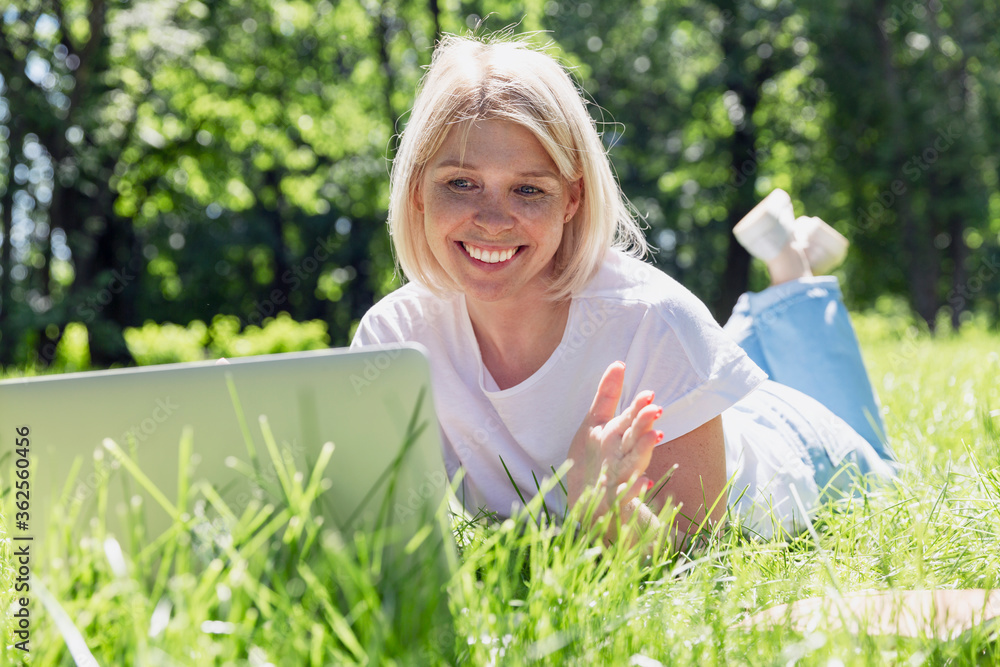 A smiling young blonde woman lies with a laptop on the grass in a park on a sunny summer day. Blogging, online communication and social networks. A smiling young blonde woman lies with a laptop on the grass in a park on a sunny summer day. Blogging, online communication and social networks.