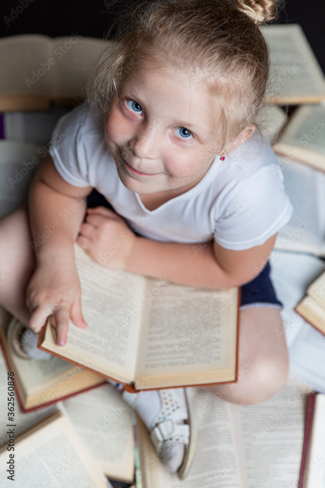 A smiling little girl is sitting on the floor with books. Education and training. Black background. Vertical. Top view. A smiling little girl is sitting on the floor with books. Education and training. Black background. Vertical. Top view.