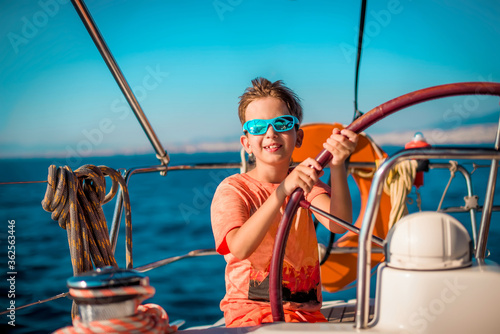 A handsome boy of 9-10 years old at the helm of a yacht in bright orange clothes, blue glasses against a blue sky. Sea vacation concept, sailing regatta. Traveling with children. photo