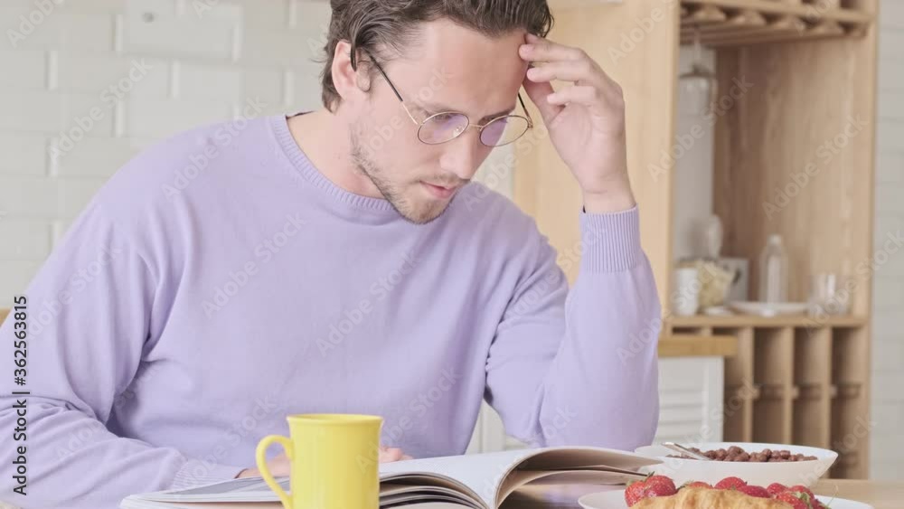 Focused handsome man in eyeglasses reading magazine while having breakfast by the table at home Focused handsome man in eyeglasses reading magazine while having breakfast by the table at home