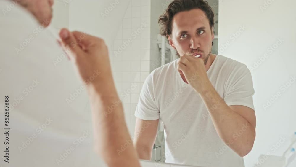 Carefree handsome man in eyeglasses brushing teeth while standing near the mirror in bathroom Carefree handsome man in eyeglasses brushing teeth while standing near the mirror in bathroom