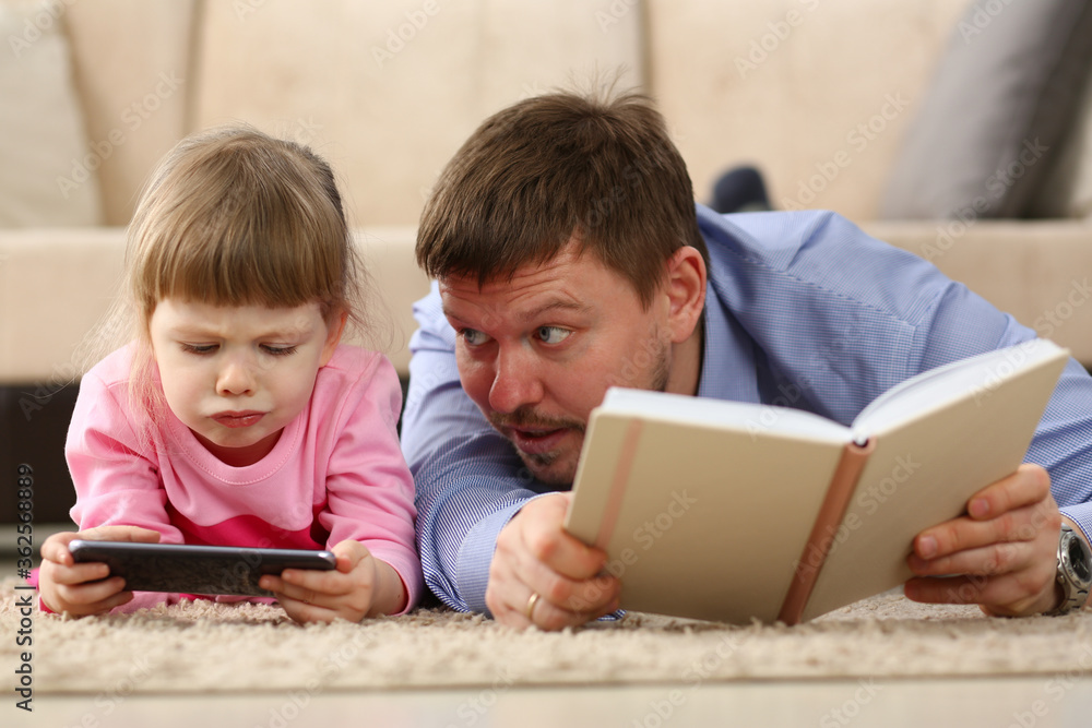 Portrait of young dad holding book and looking at her kid. Little girl using smartphone and looking at screen. Family concept Portrait of young dad holding book and looking at her kid. Little girl using smartphone and looking at screen. Family concept