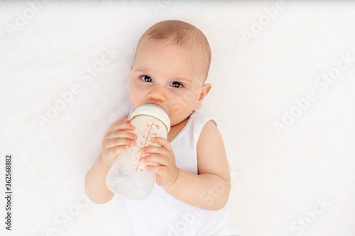 baby boy 8 months old lying on the bed in the nursery on his back and holding a bottle of milk, feeding the baby, baby food concept © Any Grant