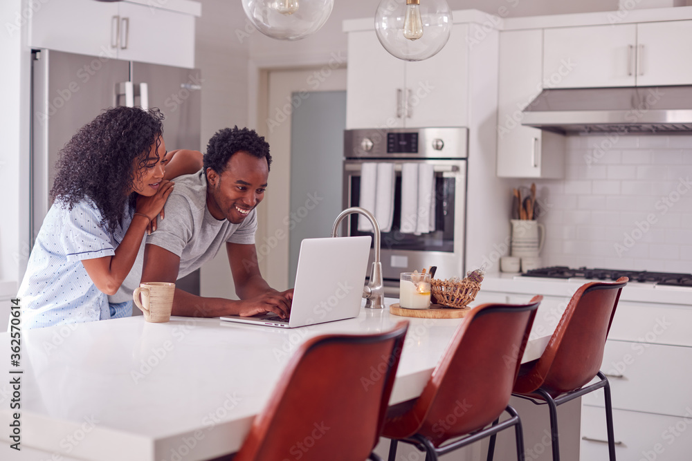Couple Wearing Pyjamas Standing In Kitchen Working From Home On Laptop Couple Wearing Pyjamas Standing In Kitchen Working From Home On Laptop