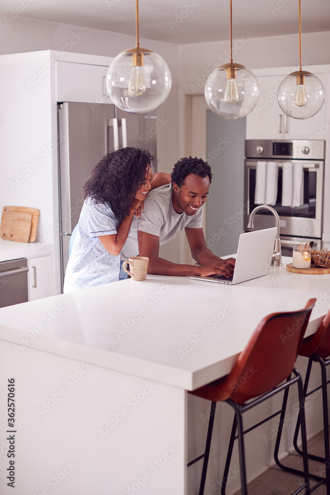 Couple Wearing Pyjamas Standing In Kitchen Working From Home On Laptop Couple Wearing Pyjamas Standing In Kitchen Working From Home On Laptop