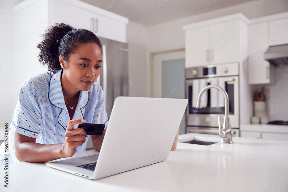 Woman Wearing Pyjamas In Kitchen Making Online Credit Card On Laptop Woman Wearing Pyjamas In Kitchen Making Online Credit Card On Laptop