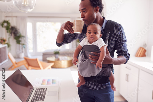 Father With Baby Daughter In Sling Multi-tasking Working From Home On Laptop