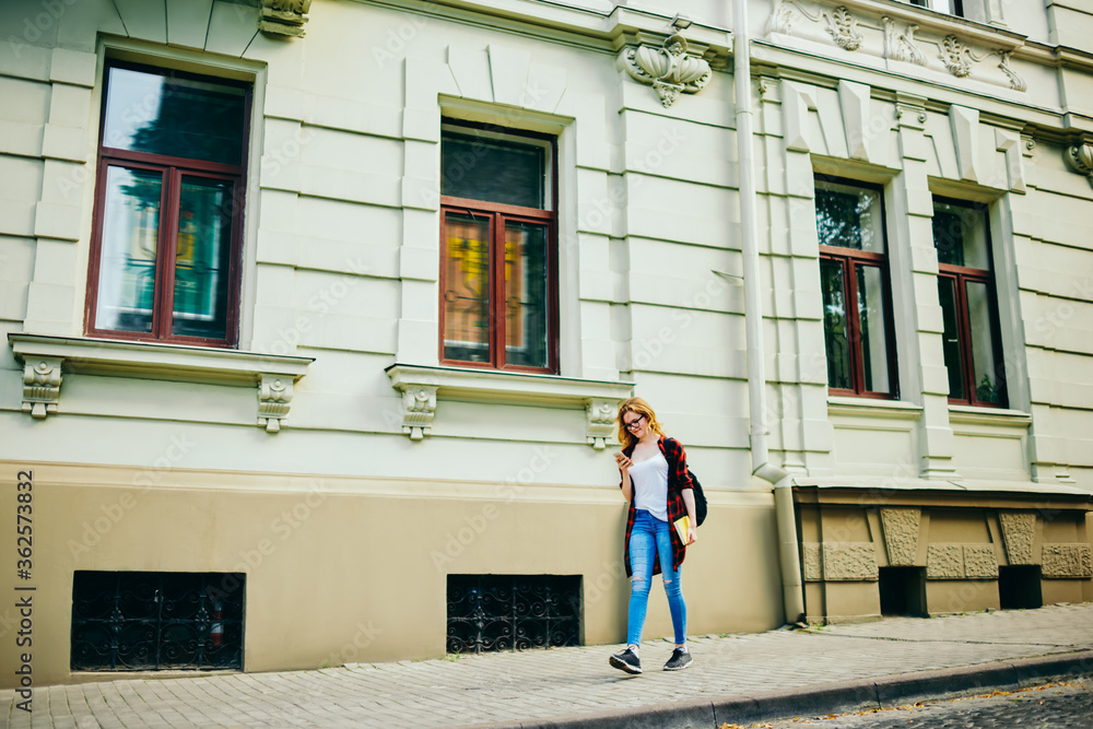 Young hipster girl walking on street passing architecture buildings on street using mobile application for searching direction,traveler in casual outfit strolling during free time on urban settings