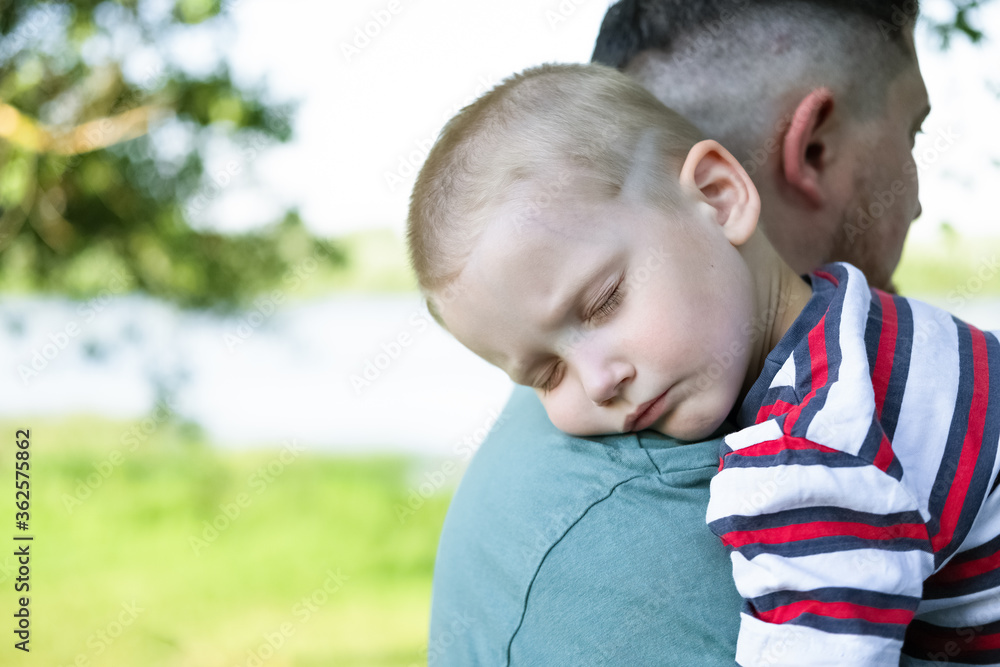 A young child is sleeping in his father's safe and protective embrace while on walk. Portrait of cute adorable blond caucasian toddler boy sleeping on fathers shoulder in park. Single father concept.