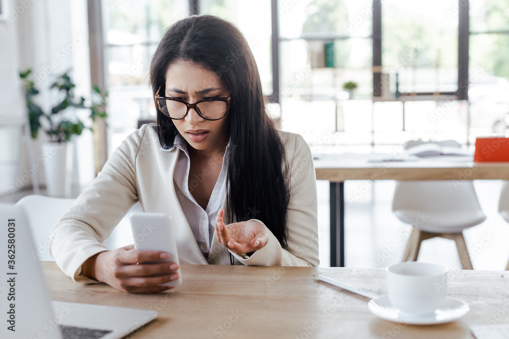 selective focus of displeased businesswoman using smartphone and gesturing near laptop and cup selective focus of displeased businesswoman using smartphone and gesturing near laptop and cup