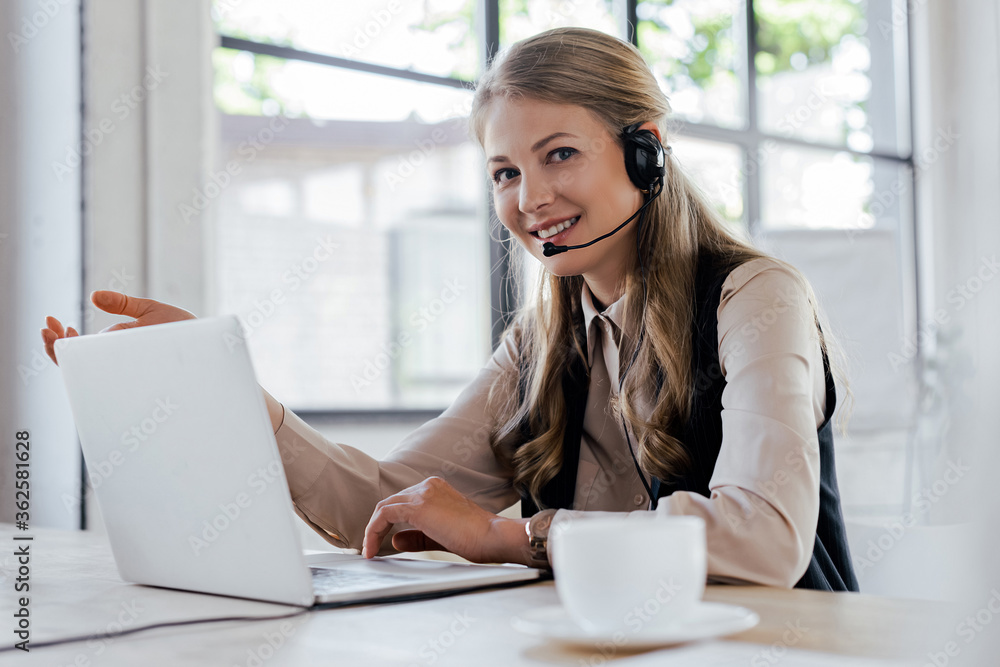 selective focus of happy operator in headset smiling near laptop and cup selective focus of happy operator in headset smiling near laptop and cup