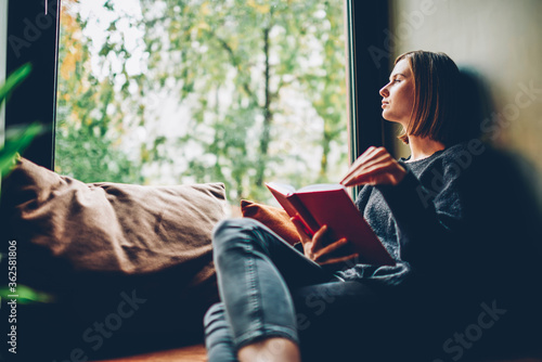 Thoughtful young woman looking out of window pondering on idea and plot of new bestseller of favourite author resting on comfortable windowsill in stylish coworking space.Copy space area