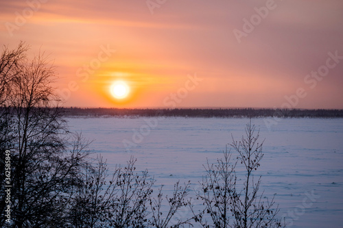 Landscape of sundown over a frozen river and snowy forest in the background.