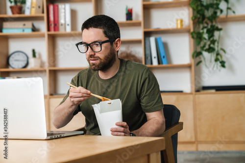 Image of man eating asian noodles takeaway while working with laptop