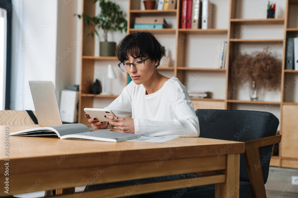 Image of focused woman in eyeglasses working with laptop and cellphone Image of focused woman in eyeglasses working with laptop and cellphone