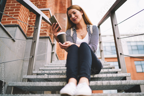 Fototapeta Naklejka Na Ścianę i Meble -  Smiling female student 20 years old watching new episode from serial via modern telephone while resting outdoors, happy hipster girl sitting at stairs enjoying coffee and messaging via mobile phone