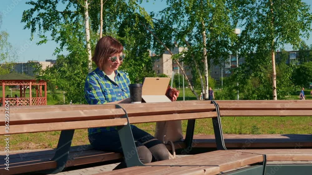 Smiling female freelancer in blue shirt does the work remotely using digital tablet in the summer park. Young woman in sunglasses sits at wooden table with sweet bun and paper cup with coffee. Smiling female freelancer in blue shirt does the work remotely using digital tablet in the summer park. Young woman in sunglasses sits at wooden table with sweet bun and paper cup with coffee.