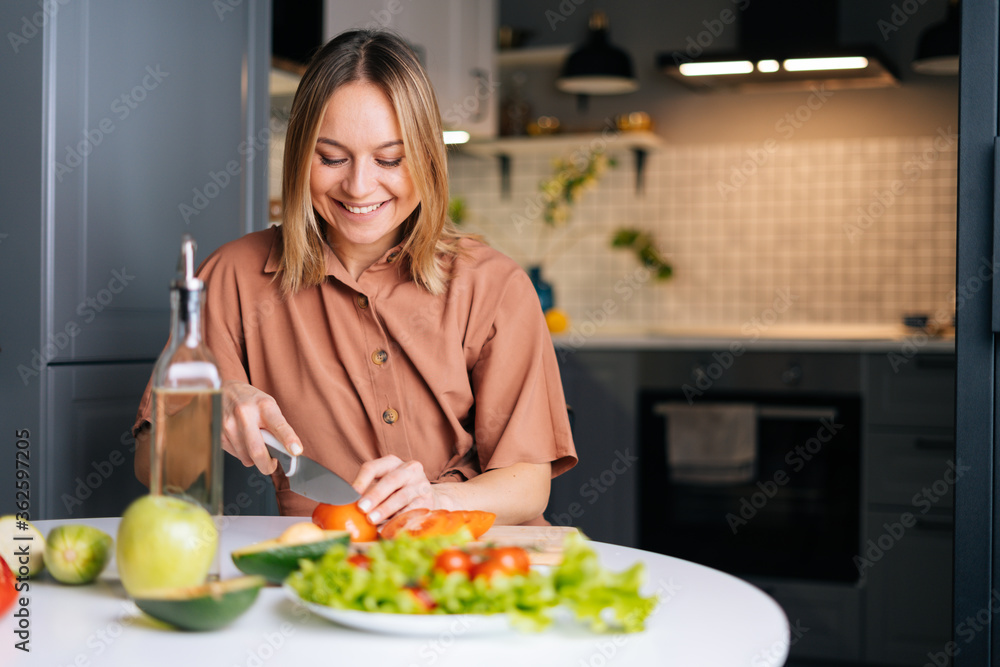 Happy smiling woman cutting fresh organic tomato with a knife for vegetable salad in modern kitchen. Smiling slim vegetarian female prepare healthy vegan dinner meal. Concept of healthy eating. Happy smiling woman cutting fresh organic tomato with a knife for vegetable salad in modern kitchen. Smiling slim vegetarian female prepare healthy vegan dinner meal. Concept of healthy eating.