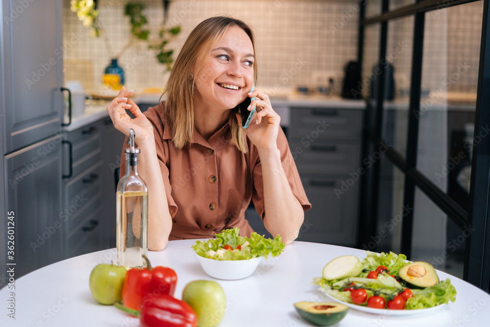 Beautiful young smiling woman talking on phone at the table with vegetables, fruits and salad in kitchen with modern interior. Concept of healthy eating.