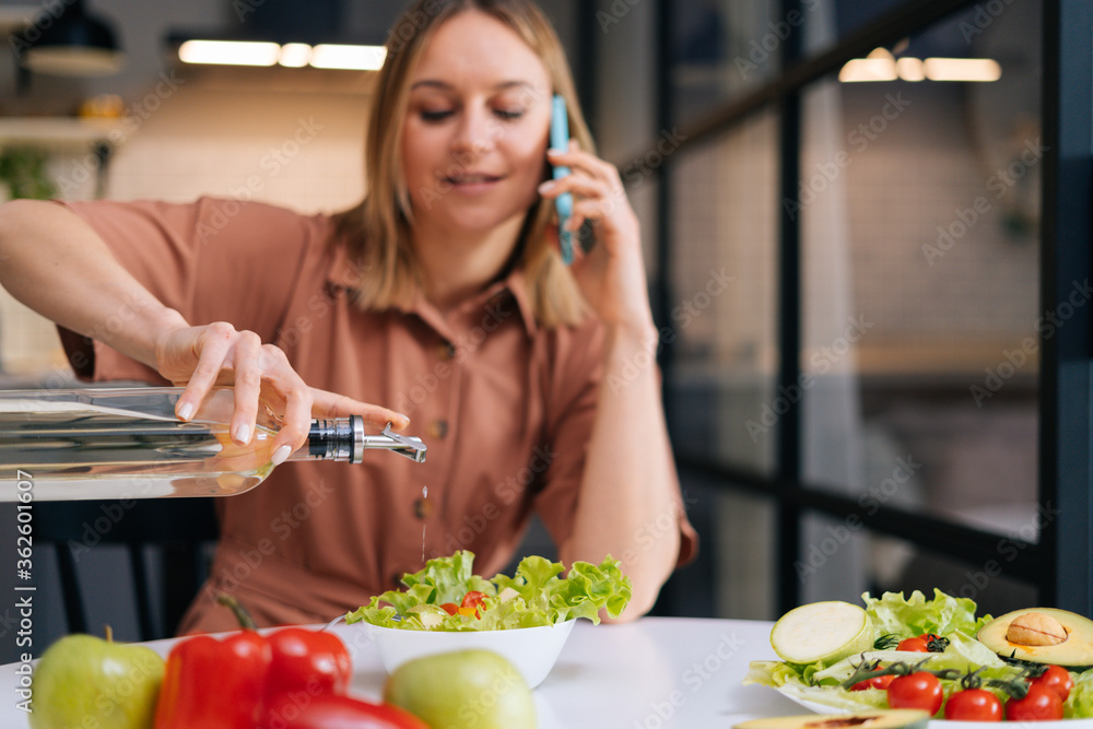 Cheerful young woman pours olive oil into vegetable salad and talks on the phone in kitchen with modern interior. Concept of healthy eating.