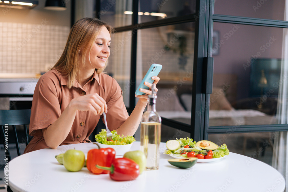 Smiling young woman using her mobile phone while eating salad in cozy wooden kitchen. Concept of healthy eating. Smiling young woman using her mobile phone while eating salad in cozy wooden kitchen. Concept of healthy eating.