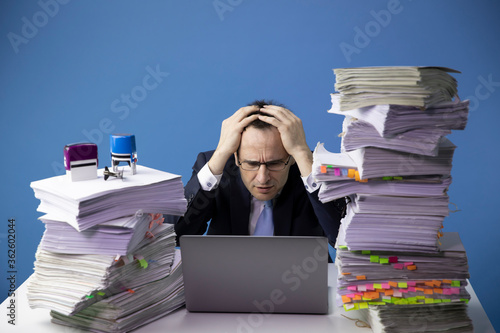 exhausted office employee working on laptop computer wrapped his arm around his head, surrounded by high stacks of documents doing overtime project isolated on blue. bureaucracy concept