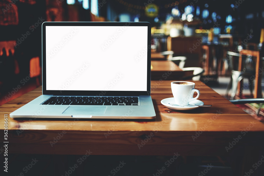 Fototapeta premium Open laptop computer and cup of coffee lying on a wooden table in cafe bar interior, portable net-book with copy space screen for your information content or text message, distance work via internet