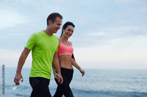 Fototapeta Naklejka Na Ścianę i Meble -  Sport couple walking along the beach resting after workout, sexy fit woman and man dressed in fluorescent-shirt taking break after run, laughing couple walk along seashore after fitness training