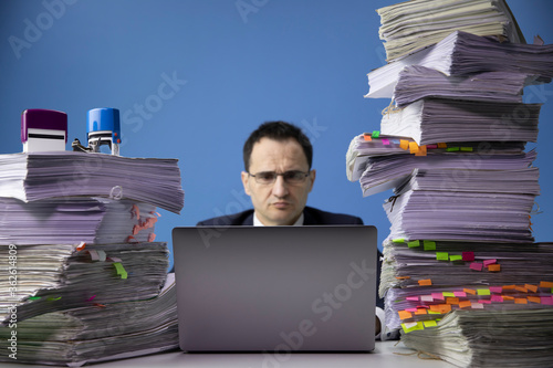 businessman works on computer laptop at office desk with huge pile of documents.