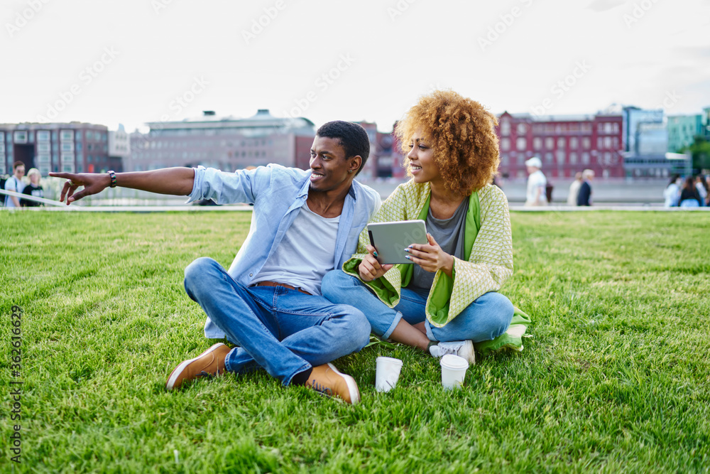 Positive dark skinned hipster guy pointing hand on side while resting with girlfriend on green grass in park.Cheerful two friends laughing while installing app on touch pad using 4G internet Positive dark skinned hipster guy pointing hand on side while resting with girlfriend on green grass in park.Cheerful two friends laughing while installing app on touch pad using 4G internet