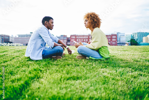 Best african american friends dressed in casual wear resting in park sitting in lotus pose on green grass and communicating with each other.Dark skinned female with smartphone talking with boyfriend