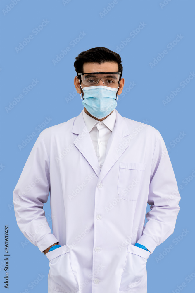 Portrait of confident male Middle Eastern Arab researcher scientist medical doctor wearing surgical mask and protection glasses standing and put hands in pockets isolated on light blue background Portrait of confident male Middle Eastern Arab researcher scientist medical doctor wearing surgical mask and protection glasses standing and put hands in pockets isolated on light blue background