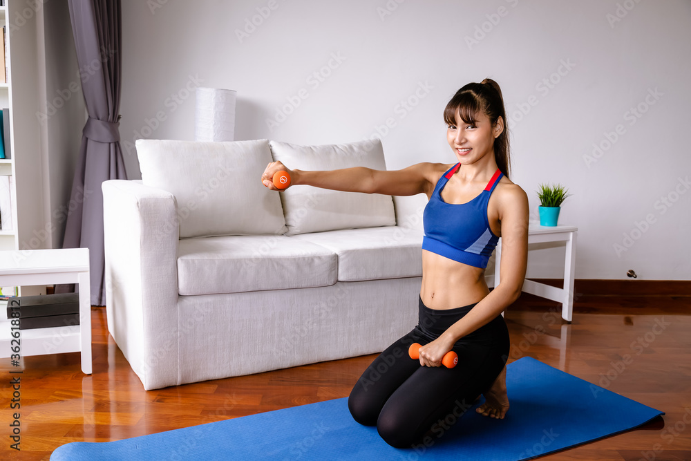 Fototapeta premium Attractive sporty woman doing dumbbell weight exercises in living room at home, sitting on floor, looking at camera