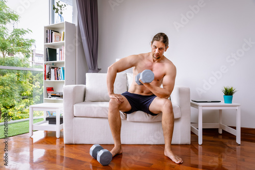 Attractive handsome muscular man doing dumbbell weight exercises in living room at home, looking at camera