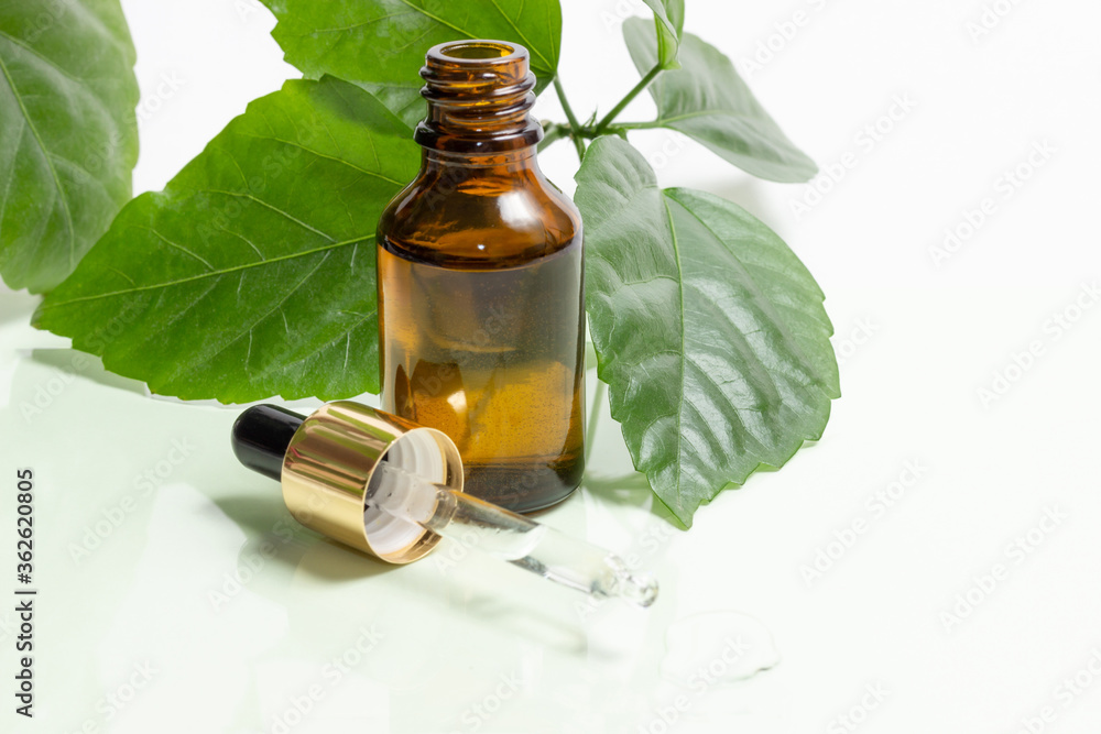 Glass cosmetic bottles with a dropper stand next to green leaves on a white background. Organic cosmetics concept, natural essential oil and cream. Glass cosmetic bottles with a dropper stand next to green leaves on a white background. Organic cosmetics concept, natural essential oil and cream.