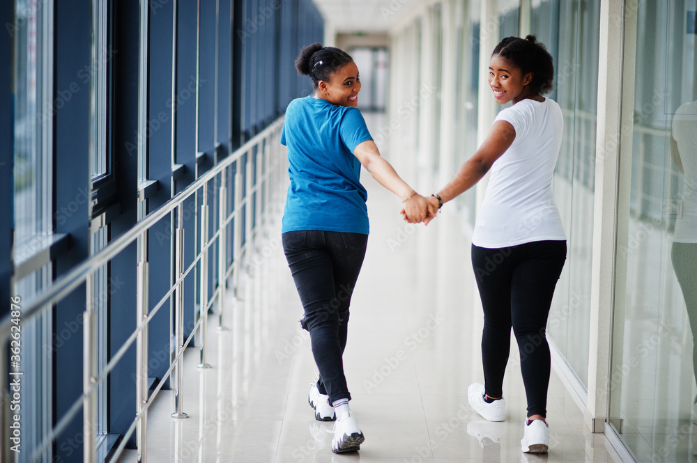 Back of two african woman friends in t-shirts walking halding hands indoor together. Back of two african woman friends in t-shirts walking halding hands indoor together.