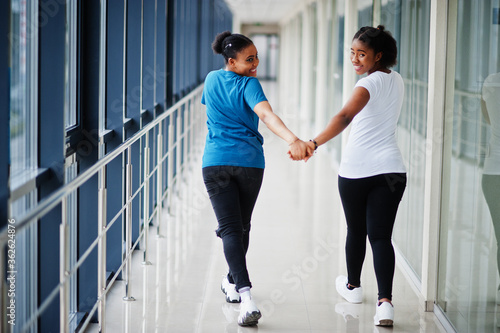 Back of two african woman friends in t-shirts walking halding hands indoor together. © AS Photo Family
