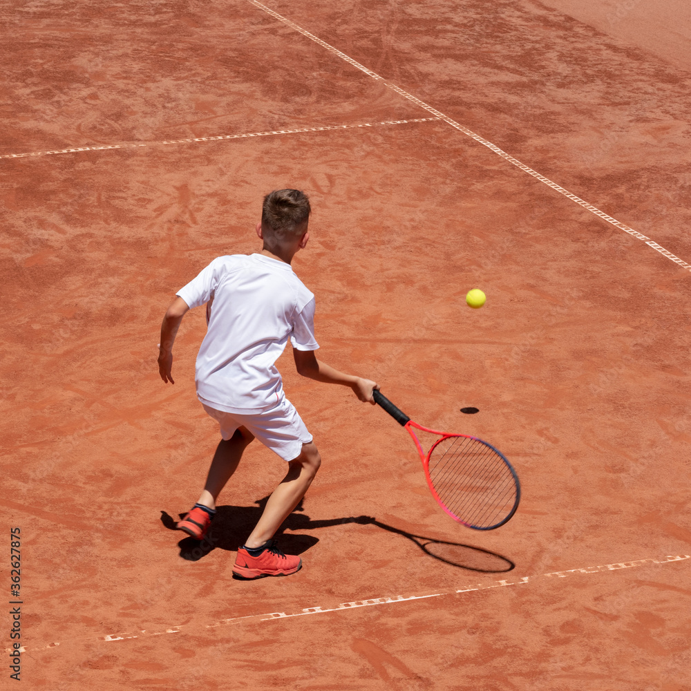 Boy plays tennis on a clay tennis court. Child is concentrated and focused on the game. Young tennis player with racket in action. Kids tennis sport concept. Motion. Shadow. Copy space