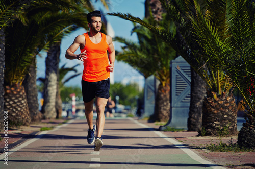 Full length portrait of muscular build athletic man jogging at sunny afternoon outdoors, handsome male jogger running down lane with palm trees on the sides and copy space area for your text message