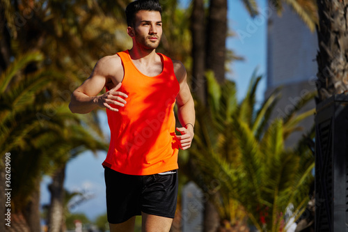 Cropped shot of handsome male runner jogging fast in tropic urban setting, muscular build man dressed in bright t-shirt with copy space area for text message or advertise content working out outdoors