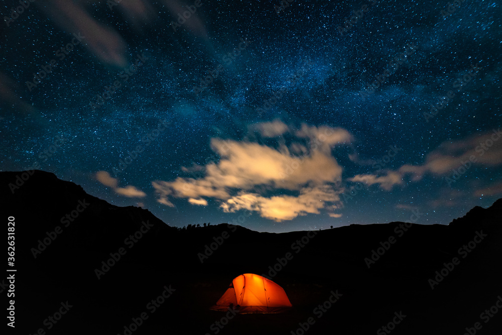 Orange tent with a burning light inside stands in a clearing under the starry sky. On the horizon mountains and forest. Concept of a beautiful starry night sky and the Milky Way. Night in the mountain